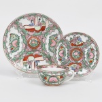 Elegante trio para chá em porcelana chinesa, decoração policromada no estilo famille-rose, esmaltação branca, apresentando decoração policromada esmaltada sobre o vidrado, predominantemente em tons de rosa, verde, vermelho, amarelo e azul, com realces em contorno escuro e filete dourado no bordo; ornamentação segue repertório decorativo inspirado no estilo famille-rose, caracterizado por composições em reservas cartuchadas delimitadas por volutas e elementos fitomórficos, reservas apresentam cenas figurativas com personagens chineses em ambiente doméstico, acompanhadas por painéis com motivos florais e frutíferos estilizados, organizada de forma radial no prato e no pires, com medalhão central decorado por motivo floral, bordo realçado por filete dourado, característica recorrente em porcelanas chinesas destinadas ao mercado de exportação. Verso com  marca em selo vermelho com caracteres chineses, indicando fabricação na China. Medidas: Xícara: Alt.: 5 x Comp.: 11,5 x Larg.: 10 cm; Pires: Diam.: 14 cm; Prato: Diam.: 19 cm.