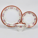 NORITAKE, Nagoya - Japão(Fundada em 1904 pelos irmãos Ichizaemon e Toyo Morimura). Exuberante chávena para chá executada em porcelana japonesa, esmaltagem vítrea na cor branca, circundada por buquês de flores em estampagem policromada sobre fundo levemente em tom marfim, dotada de profusa ornamentação em douradura, com frisos, arabescos, e elementos vegetalistas sobrepondo faixa de tonalidade bordô contornando as bordas, bojo campaniforme sobre base circular, alça lateral em voluta, padrão decorativo replicado no pires. Marca da manufatura ao fundo. Meds.: Chávena: Alt. 6 x Comp. 12 x Larg. 9,5 cm. Pires: Diam. 14,5 cm. Prato: Diam.: 19 cm. Apresenta desgastes na douradura, vide foto.