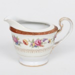 NORITAKE, Nagoya - Japão(Fundada em 1904 pelos irmãos Ichizaemon e Toyo Morimura). Exuberante cremeira executada em porcelana japonesa, esmaltagem vítrea na cor branca, circundado por buquês de flores em estampagem policromada sobre faixa levemente em tom marfim, dotada de profusa ornamentação em douradura, com frisos, arabescos, e elementos vegetalistas sobrepondo faixa de tonalidade bordô contornando a borda, borda ondulada e revirada para fora, alça lateral em voluta. Marca da manufatura ao fundo. Med.: Alt. 10 x Comp. 15 x Larg. 10 cm.