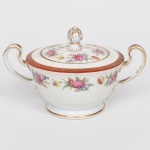 NORITAKE, Nagoya - Japão(Fundada em 1904 pelos irmãos Ichizaemon e Toyo Morimura). Exuberante açucareiro executado em porcelana japonesa, esmaltagem vítrea na cor branca, circundado por buquês de flores em estampagem policromada sobre faixa levemente em tom marfim, dotada de profusa ornamentação em douradura, com frisos, arabescos, e elementos vegetalistas sobrepondo faixa de tonalidade bordô contornando a borda, tampa encimada por puxador modelado em forma de fruto, bico curvilíneo de ponta recortada, alças laterais em oluta. Marca da manufatura ao fundo. Med.: Alt. 11,5 x Comp. 18,5 x Larg. 13 cm.