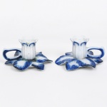 Par de pequenos castiçais em porcelana moldada em forma de flor estilizada, com base em pétalas abertas decoradas em azul cobalto sobre fundo branco. Cada peça apresenta haste central estriada com borda em forma de corola e alça lateral vazada para manuseio. No verso consta a marca Porcelana Monte Sião - MG. Medidas: Alt.: 6 x Comp.: 11 x Larg.: 9,5 cm. Uma apresenta um ínfimo bicado na borda da bobeche.