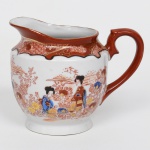 Leiteira executada em porcelana japonesa decorada em esmaltes policromos no estilo Imari, esmaltação branca, corpo globular assente sobre base circular, provida de bico vertedor ondulado e alça lateral em voluta, esta integralmente esmaltada em vermelho-ferro; decoração em esmaltes policromos sobre o vidrado, predominando a tonalidade vermelho-ferro, complementado por azul, amarelo, verde e douração, bojo com cenas figurativas com personagens femininas em trajes tradicionais japoneses, representadas em meio a paisagem estilizada com arquitetura, vegetação e composição floral abundante, parte superior ornada por faixa decorativa recortada com reservas geométricas e florais, com contornos realçados em preto e detalhes em dourado, característica comum da porcelana japonesa inspirada no repertório ornamental Imari, face oposta apresenta paisagem estilizada com construções e vegetação, executada em traço monocromático vermelho, complementada por flores policromas na parte inferior da composição. Fundo com marca impressa em vermelho MADE IN JAPAN, indicando produção destinada ao mercado de exportação. Medidas: Alt.: 9,5 x Comp.: 12 x Larg.: 8,5 cm.