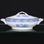 ROSSLAU - ALEMANHA. Exuberante terrina com tampa oval em porcelana alemã, tampa abaulada encimada por puxador em arco, alças laterais reviradas para cima, relevadas com volutas na própria pasta. Esmaltação branca, decoração apresenta composição em azul padrão china-blau, característica da tradição decorativa centro-europeia inspirada nas porcelanas orientais; aba ornada por friso contínuo em azul com motivos florais estilizados e elementos geométricos, delimitado por filete dourado acompanhando o contorno ondulado da borda, campo central apresenta vinheta decorativa em azul, composta por arranjo floral acompanhado de pequeno elemento ornamental representando ânfora sobre base. Marca na base em azul: CHINA-BLAU / ROSSLAU, indicando manufatura da fábrica Rosslau, Alemanha. Medidas: Alt.: 12 x Comp.: 33 x Larg.: 19 cm.
