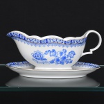 ROSSLAU - ALEMANHA. Exuberante molheira em porcelana alemã, formato navicular com vertedouro proeminente e alça lateral em voluta estilizada, contorno recortado em arcos, fixada sobre presentoir oval. Esmaltação branca, decoração apresenta composição em azul padrão china-blau, característica da tradição decorativa centro-europeia inspirada nas porcelanas orientais; aba ornada por friso contínuo em azul com motivos florais estilizados e elementos geométricos, delimitado por filete dourado acompanhando o contorno ondulado da borda, face lateral apresenta vinheta decorativa em azul, composta por arranjo floral acompanhado de pequeno elemento ornamental representando ânfora sobre base, padrão decorativo replicado no presentoir. Marca na base em azul: CHINA-BLAU / ROSSLAU, indicando manufatura da fábrica Rosslau, Alemanha. Medidas: Alt.: 9x  Comp.: 20 x Larg.: 12 cm.