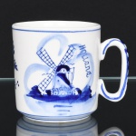 Caneca em porcelana branca, de formato cilíndrico, com base plana e bordo reto, provida de alça lateral vazada de formato oval. Decoração em azul sob esmalte, composta por cena com moinho de vento e inscrição Holland, acompanhada por elementos fitomorfos estilizados no verso, executados em pinceladas livres e sombreadas. Filetes azuis junto ao bordo e à base. Marca da manufatura no verso. Medidas: Alt.: 7,5 x Comp.: 9,5 x Larg.: 7 cm.