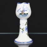 Castiçal executada em cerâmica esmaltada, Delft Holland, decorada em policromia sob vidrado, apresentando repertório ornamental inspirado na tradição cerâmica da cidade de Delft, nos Países Baixos. Bojo modelado em forma de tulipa com leve estriamento vertical e borda ondulada, apoiado sobre haste cilíndrica afunilada e base circular; ornamentado com paisagem holandesa estilizada, destacando moinho de vento à beira dágua, acompanhado de pequenas construções rurais, árvores e aves em voo, com a inscrição Holland, executada em azul cobalto, verde e amarelo, remetendo às composições figurativas associadas ao chamado Delftware, haste apresenta elementos florais estilizados e motivos pontilhados, pintados em azul e verde, compondo conjunto decorativo, filetes azuis delimitam a borda e a transição entre bojo e haste. Verso com marca impressa Delft Blue Handpainted Designed by DSM  Holland, indicando produção holandesa com decoração pintada à mão. Medidas: Alt.: 16 x Diam.: 7,5 cm.
