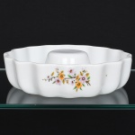Porcelana Schmidt - Brasil(Fundada em 1945). Forma tipo pudim / bolo executado em porcelana refratária, linha Calorama, produzida para uso em forno; formato circular com paredes onduladas, tubo central elevado, esmaltação branca, ornamentada com pequenos ramos florais em estampagem policromada, composto por flores em tons de amarelo, laranja e rosa, acompanhada por folhas verdes. Marca da manufatura no verso, acompanhada das inscrição "A porcelana que vai ao forno" e numeração do modelo 142/21, identificando a série de produção da manufatura. Medidas: Alt.: 5,5 x Diam.: 22 cm.