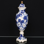 Cerâmica Irmãos Weiss, São José dos Campos - SP.(Fundada em 1940, ativa até 1995). Vaso com tampa em faiança nacional da década de 1940/50, de corpo oval alongado, assentado sobre haste torneada e base moldada de contorno recortado. Bojo com decoração floral em azul sob esmaltação branca, composta por grandes flores estilizadas e folhagem, distribuídas de forma contínua, realçada por aplicação de filetes e coberturas em douradura. Pescoço levemente constrito, abrindo em colo e borda saliente. Tampa campaniforme, com encaixe interno, rematada por pegador ovoide. Marca da manufatura ao fundo. Apresenta quebradura colada na borda, ínfimo trincado na borda de encaixe da tampa, e um pequeno desgaste da esmaltação no bojo. Medidas: Altura: 46 x Diâmetro: 14,5 cm.