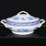 ROSSLAU - ALEMANHA. Exuberante sopeira oval em porcelana alemã, tampa abaulada encimada por puxador em arco, dotada de recorte lateral (entaille) destinado à passagem da concha de serviço, alças laterais reviradas para cima, relevadas com volutas na própria pasta. Esmaltação branca, decoração apresenta composição em azul padrão china-blau, característica da tradição decorativa centro-europeia inspirada nas porcelanas orientais; aba ornada por friso contínuo em azul com motivos florais estilizados e elementos geométricos, delimitado por filete dourado acompanhando o contorno ondulado da borda, campo central apresenta vinheta decorativa em azul, composta por arranjo floral acompanhado de pequeno elemento ornamental representando ânfora sobre base. Marca na base em azul: CHINA-BLAU / ROSSLAU, indicando manufatura da fábrica Rosslau, Alemanha. Medidas: Alt.: 18 x Comp.: 38 x Larg.: 22 cm.