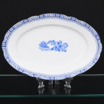 ROSSLAU - ALEMANHA. Exuberante travessa oval em porcelana alemã, de contorno recortado com aba larga levemente ondulada. Esmaltação branca, decoração apresenta composição em azul padrão china-blau, característica da tradição decorativa centro-europeia inspirada nas porcelanas orientais; aba ornada por friso contínuo em azul com motivos florais estilizados e elementos geométricos, delimitado por filete dourado acompanhando o contorno ondulado da borda, campo central apresenta vinheta decorativa em azul, composta por arranjo floral acompanhado de pequeno elemento ornamental representando ânfora sobre base. Marca na base em azul: CHINA-BLAU / ROSSLAU, indicando manufatura da fábrica Rosslau, Alemanha. Medidas: Comp.: 29,5 x Larg.: 19,5 cm.