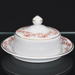 Porcelana Rio Branco/Cia. Brasileira de Vidros, Rio de Janeiro - RJ(Fundada em 1947, ativa até 1967). Elegante manteigueira executada em porcelana nacional da década de 1950/50; esmaltagem vítrea na cor branca, decoração em esmalte vermelho-coral, formada por arabescos e ramos vegetalistas estilizados distribuídos ao longo da borda da tampa e da aba do prato, realçada com filetes em douradura; tampa encimada por puxador oviforme, presentoir de centro rebaixado destinado ao encaixe da manteigueira. Marca da manufatura no verso. Medidas: Alt.: 8 x Diam.: 14,5 cm.