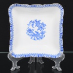 ROSSLAU - ALEMANHA. Exuberante saladeira em porcelana alemã, formato quadrangular, de quinas arredondadas, contorno recortado com aba larga e ondulada. Esmaltação branca, decoração apresenta composição em azul padrão china-blau, característica da tradição decorativa centro-europeia inspirada nas porcelanas orientais; aba ornada por friso contínuo em azul com motivos florais estilizados e elementos geométricos, delimitado por filete dourado acompanhando o contorno ondulado da borda, campo central apresenta vinheta decorativa em azul, composta por arranjo floral acompanhado de pequeno elemento ornamental representando ânfora sobre base. Marca na base em azul: CHINA-BLAU / ROSSLAU, indicando manufatura da fábrica Rosslau, Alemanha. Medidas: Alt.: 6 x Comp.: 19 x Larg.: 19 cm.