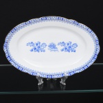 ROSSLAU - ALEMANHA. Exuberante travessa oval em porcelana alemã, de contorno recortado com aba larga levemente ondulada. Esmaltação branca, decoração apresenta composição em azul padrão china-blau, característica da tradição decorativa centro-europeia inspirada nas porcelanas orientais; aba ornada por friso contínuo em azul com motivos florais estilizados e elementos geométricos, delimitado por filete dourado acompanhando o contorno ondulado da borda, campo central apresenta vinheta decorativa em azul, composta por arranjo floral acompanhado de pequeno elemento ornamental representando ânfora sobre base. Marca na base em azul: CHINA-BLAU / ROSSLAU, indicando manufatura da fábrica Rosslau, Alemanha. Medidas: Comp.: 29,5 x Larg.: 19,5 cm.