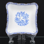 ROSSLAU - ALEMANHA. Exuberante saladeira em porcelana alemã, formato quadrangular, de quinas arredondadas, contorno recortado com aba larga e ondulada. Esmaltação branca, decoração apresenta composição em azul padrão china-blau, característica da tradição decorativa centro-europeia inspirada nas porcelanas orientais; aba ornada por friso contínuo em azul com motivos florais estilizados e elementos geométricos, delimitado por filete dourado acompanhando o contorno ondulado da borda, campo central apresenta vinheta decorativa em azul, composta por arranjo floral acompanhado de pequeno elemento ornamental representando ânfora sobre base. Marca na base em azul: CHINA-BLAU / ROSSLAU, indicando manufatura da fábrica Rosslau, Alemanha. Medidas: Alt.: 6,5 x Comp.: 21 x Larg.: 21 cm.