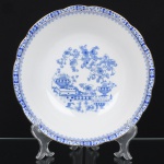 ROSSLAU - ALEMANHA. Exuberante bowl em porcelana alemã, formato hemisférico, contorno recortado e borda ligeiramente revirada para fora. Esmaltação branca, decoração apresenta composição em azul padrão china-blau, característica da tradição decorativa centro-europeia inspirada nas porcelanas orientais; aba ornada por friso contínuo em azul com motivos florais estilizados e elementos geométricos, delimitado por filete dourado acompanhando o contorno da borda, campo central apresenta vinheta decorativa em azul, composta por arranjo floral acompanhado de pequeno elemento ornamental representando ânfora sobre base. Marca na base em azul: CHINA-BLAU / ROSSLAU, indicando manufatura da fábrica Rosslau, Alemanha. Medidas: Alt.: 6,5 x Diam.: 26 cm.