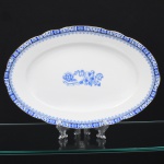 ROSSLAU - ALEMANHA. Exuberante travessa oval em porcelana alemã, de contorno recortado com aba larga levemente ondulada. Esmaltação branca, decoração apresenta composição em azul padrão china-blau, característica da tradição decorativa centro-europeia inspirada nas porcelanas orientais; aba ornada por friso contínuo em azul com motivos florais estilizados e elementos geométricos, delimitado por filete dourado acompanhando o contorno ondulado da borda, campo central apresenta vinheta decorativa em azul, composta por arranjo floral acompanhado de pequeno elemento ornamental representando ânfora sobre base. Marca na base em azul: CHINA-BLAU / ROSSLAU, indicando manufatura da fábrica Rosslau, Alemanha. Medidas: Comp.: 33 x Larg.: 22 cm.