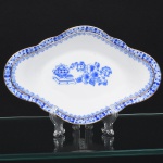 ROSSLAU - ALEMANHA. Exuberante travessa oblonga em porcelana alemã, de contorno recortado com aba larga e ondulada, interior côncavo. Esmaltação branca, decoração apresenta composição em azul padrão china-blau, característica da tradição decorativa centro-europeia inspirada nas porcelanas orientais; aba ornada por friso contínuo em azul com motivos florais estilizados e elementos geométricos, delimitado por filete dourado acompanhando o contorno ondulado da borda, campo central apresenta vinheta decorativa em azul, composta por arranjo floral acompanhado de pequeno elemento ornamental representando ânfora sobre base. Marca na base em azul: CHINA-BLAU / ROSSLAU, indicando manufatura da fábrica Rosslau, Alemanha. Medida: Alt.: 4 x Comp.: 22 x Larg.: 14 cm.
