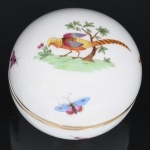 Herend Porcelain Manufactory, Herend - Hungria. Caixa circular em porcelana branca húngara, esmaltagem vítrea na cor branca, formato globular, tampa dotada de decoração manual em policromia, com cena naturalista representando ave de plumagem colorida, posicionada sobre terreno gramado junto a pequena árvore, acompanhada por borboletas multicores distribuídas ao redor da composição, bordas contornadas por friso em douradura. Verso apresenta marca azul com inscrição Handpainted  Herend  Hungary, acompanhada do brasão da manufatura, indicando peça pintada à mão. Medidas: Alt.: 7,5 x Diam.: 9,5 cm.