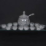 Conjunto de poncheira em cristal incolor lapidado, composto por recipiente central com tampa e colher de servir, acompanhado por doze(12) xícaras com alça, destinado ao serviço de ponche ou bebidas frias; corpo globular com tampa encaixada e pomo facetado, decorado com lapidação profunda composta por estrelas, raios e facetas distribuídas ao longo da superfície, colher possui cabo curvo em cristal transparente, adaptado para o serviço da bebida, xícaras apresentam forma arredondada alça circular, igualmente ornamentadas com motivos lapidados em padrão estrelado e raiado, criando efeitos de brilho e refração da luz. Medidas: Poncheira: Alt.: 31,5 x Diam.: 24,5 cm; Concha: Comp.: 37 cm; Caneca: Alt.: 6 x Comp.: 11 x Larg.: 7,5 cm. Canecas apresentam pequenos bicados na borda. Borda da poncheira com um ínfimo bicado.