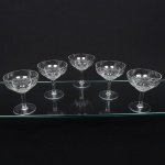 Conjunto de taças de pé alto, para champanhe, em meio cristal, diáfano e incolor; bojo hemisférico talhado com sucessão de sulcos entrecruzados formando padrão geométrico, fuste tubular sobre base circular. Composto de cinco(05) unidades. Medidas: Alt.: x Diam.: