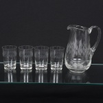 Elegante conjunto em cristal, diáfano e incolor; composto de uma(01) jarra e quatro(04) copos, lapidados com sucessão de navetes dispostas horizontalmente, jarra de bojo balaústre, bico vertedor e borda ondulados, alça lateral em voluta aplicada sobre a superfície, copos cilíndricos de base espessa. Medidas: Jarra: Alt.: x Comp.: x Larg.: ; Copos: Alt.: x Diam.: