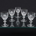 Conjunto de taças em espesso cristal, diáfano e incolor; bojo oval circundado por facetas verticais formando arcos, haste hexagonal com nó intermediário apoiado sobre base circular. Padrão similar ao "harcourt", da manufatura Baccarat. Composto de cinco(05) unidades. Medidas: Alt.: x Diam.: