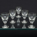 Conjunto composto por cinco(05) cálices em meio cristal incolor, apresentando bojo ovóide levemente afunilado, apoiado sobre haste curta cilíndrica com anel central e base circular plana. Medidas: Alt.: x Diam.: