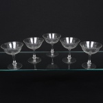 Conjunto de taças de pé alto para champanhe, executado em cristal incolor; bojo hemisférico, talhado com decoração geométrica composta por linhas entrecruzadas, intercalado por motivos radiais em forma de estrela ou palmeta, haste alta torneada com leve dilatação central e base circular plana. Composto de cinco(05) unidades. Medidas: Alt.: x Diam.: