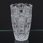 Vaso executado em cristal incolor espesso, prensado e moldado, de corpo alto e facetado, apresentando perfil poligonal com painéis verticais profundamente lapidados, bordo recortado em ondulações lobadas, acompanhando o ritmo das faces do corpo; decoração geométrica profunda, composta por rosáceas radiais, estrelas multifacetadas, painéis ovais, cruzamentos de losangos e texturas pontilhadas em padrão diamante, organizadas em campos verticais alternados, base apresenta grande rosácea estrelada lapidada, típica de peças de cristal decorativo. Medidas: Alt.: x Diam.: