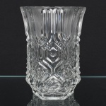 Vidro incolor moldado e prensado, corpo de secção hexagonal, com paredes facetadas e ligeira expansão superior. Borda recortada acompanhando a geometria hexagonal do corpo. Base poligonal com fundo plano; decoração obtida por moldagem em relevo, composta por registros geométricos verticais alternando painéis estriados e campos com motivos losangulares concêntricos e formas angulares escalonadas, zona superior apresenta estrias verticais paralelas, enquanto a zona média e inferior desenvolve composição geométrica simétrica estruturada por eixos verticais e diagonais. Medidas: Alt.: Diam.: