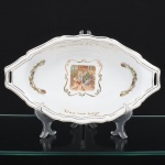 Travessa em porcelana, formato oval com extremidades recortadas e alças vazadas integradas, borda ondulada com filetes em ouro. Decoração policromada com cena figurativa central de caráter campestre, enquadrada por volutas em douração; reservas laterais com elementos fitomorfos; inscrição O pão nosso de cada dia na aba. Medidas: