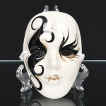 Máscara estilo veneziano de rosto feminino estilizado, modelada em cerâmica esmaltada e decorada em preto, branco e dourado, com maquiagem dramática e motivos gráficos curvilíneos que percorrem o rosto. Apresenta orifícios laterais para fixação, indicando uso como ornamento de parede. Medidas: