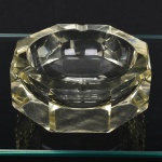 Elegante cinzeiro em cristal, diáfano, de leve tonalidade âmbar, formato octogonal, com cavidade central circular e entalhe na borda para apoio de cigarro, paredes externas apresentam facetas geométricas, produzindo efeito de refração da luz. Medidas: