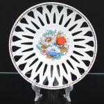 Fruteira em porcelana nacional, esmaltação branca vitrificada, formato circular com paredes vazadas em recortes geométricos, formando composição radial, centro ornamentado por arranjo floral em estampagem policromada, composto por flores estilizadas em tons de vermelho, azul, amarelo e verde, borda realçada por friso decorativo com pequenos elementos florais e geométricos, complementando a ornamentação central. Marca da manufatura no verso. Medidas: Alt.: x Diam.: