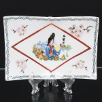 Travessa em porcelana esmaltada, tonalidade branca, forma retangular alongada com bordos ondulados, decoração em estampagem policromada, com cartela losangular central contendo figura feminina oriental, paisagem com pagode e ponte, inscrição em caracteres chineses e ramagens florais na aba. Friso geométrico linear em verde contornando o bordo. Marca da manufatura no verso. Medidas: Alt.: x Comp.: x Larg.: