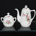 Porcelana Real, Mauá - SP(Fundada em 1943 por Fritz Erwin Schmidt). Um(01) bule para café e um açucareiro executados em porcelana da década de 1950; esmaltação branca vitrificada, bojo alto de perfil balaústre, com bico vertedor curvilíneo e alça lateral moldada em voluta, acompanhada de tampa com pegador central. O açucareiro possui bojo globular com duas pequenas alças laterais e tampa ajustada com botão superior; decoração composta por motivos florais policromados com rosas em tonalidades de rosa e folhagens em verde, aplicados sobre o esmalte branco em composição central nos corpos das peças e repetidos nas tampas, realçado com filetes em verde nos bordos e no bico vertedor. Marca da manufatura no verso. Medidas: