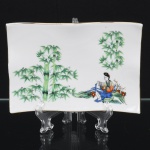 Travessa executada em porcelana branca vitrificada, de formato retangular com bordas onduladas e perfil sinuoso, apoiada sobre base plana integrada, modelagem apresenta desenho elegante e levemente curvado, característico de peças destinadas tanto ao serviço de mesa quanto ao uso decorativo; a decoração apresenta composição oriental policromada, predominando as tonalidades  verde, azul, vermelho e preto aplicadas sobre o esmalte branco, representando figura masculina em traje tradicional sentado em paisagem com bambus, acompanhada por vegetação estilizada e elementos naturais, borda contornada por friso em douradura. Marca da manufatura no verso. Medidas: Alt.: x Comp.: x Larg.: