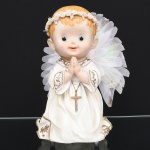Escultura em resina moldada e policromada, representando anjo infantil em gesto de oração, mãos unidas junto ao peito, portando cruz pendente. Traja veste longa com aplicação de elementos decorativos em relevo e douração. Asas posteriores em tecido com acabamento iridescente. Medidas: