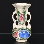 Vaso executado em cerâmica esmaltada, corpo bojudo com colo alongado e boca alargada de borda ondulada. Apresenta duas alças laterais vazadas, integradas ao corpo. Decoração floral aplicada à mão, com composição de flores em policromia (tons de azul, rosa e verde) sobre fundo claro, com traços livres e áreas de preenchimento irregular. Base circular integrada.Estado de conservação conforme fotografias, com marcas de uso; desgastes no esmalte, especialmente na borda e nas arestas. Medidas:
