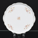 Porcelana Renner, Porto Alegre - RS(Fundada em 1937). Bandeja circular em porcelana nacional, esmaltação branca, aba larga e borda recortada em contorno ondulado. Decoração por decalque com motivos florais distribuídos na aba e no centro, em policromia (tons de rosa, amarelo e verde), realçado com filete dourado aplicado na borda. Marca da manufatura no verso. Medidas: Diam.