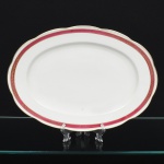 KPM - Krister Porzellan-Manufaktur, Waldenburg - Alemanha(Fundada em 1831 por Karl Kristen). Travessa oval em porcelana polonesa, esmaltação branca, com aba levemente ondulada. Decoração em faixa contínua junto à borda, composta por fundo em tom magenta com motivo de folhagem estilizada em douração, delimitada por filetes dourados. Marca de manufatura no verso. Medida: Comp.: x Larg.: cm.