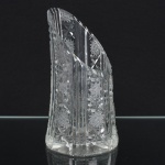 Vaso executado em cristal lapidado, de corpo alto e assimétrico, com borda superior em corte oblíquo. Decoração executada por lapidação profunda, composta por painéis verticais alternando caneluras e reservas com motivos geométricos, incluindo rosetas radiais, estrelas multifacetadas e elementos prismáticos dispostos em repetição. Base circular espessa, com lapidação inferior em desenho radial. Medidas: