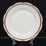 KPM - Krister Porzellan-Manufaktur, Waldenburg - Alemanha(Fundada em 1831 por Karl Kristen). Prato de serviço em porcelana polonesa, esmaltação branca, com aba levemente ondulada. Decoração em faixa contínua junto à borda, composta por fundo em tom magenta com motivo de folhagem estilizada em douração, delimitada por filetes dourados. Marca de manufatura no verso. Medida: Diam.: