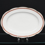 KPM - Krister Porzellan-Manufaktur, Waldenburg - Alemanha(Fundada em 1831 por Karl Kristen). Travessa oval em porcelana polonesa, esmaltação branca, com aba levemente ondulada. Decoração em faixa contínua junto à borda, composta por fundo em tom magenta com motivo de folhagem estilizada em douração, delimitada por filetes dourados. Marca de manufatura no verso. Medida: Comp.: x Larg.: cm.