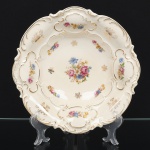 Centro de mesa em porcelana Bavaria (Alemanha), corpo circular de borda recortada e ondulada, interior côncavo, com relevo moldado de inspiração rococó. Decoração em esmaltação policroma com arranjos florais distribuídos em reservas, centro com buquê floral, e realces em douração nos filetes e elementos em relevo. Marca na base em esmalte verde sob vidrado Bavaria  Made in Germany. Medidas: