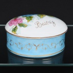Caixa com tampa em porcelana, formato oval com tampa abaulada. Decoração policromada com composição floral pintada à mão na tampa, em tons de azul, rosa e verde. Corpo com faixa em fundo azul, ornada por friso contínuo em douração. Inscrição Beatriz aplicada em douração na tampa. Borda com filetes dourados. Medidas: