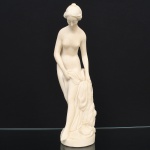 Escultura executada em marmolina moldada, representando Vênus, inspirada no modelo Vênus saindo do banho (La Baigneuse), de tradição neoclássica francesa, com referência a composições de Étienne-Maurice Falconet. Figura em posição sinuosa, com inclinação da cabeça e olhar voltado para baixo, segurando panejamento junto ao corpo. Tratamento anatômico com modelagem contínua e superfície fosca. Base circular integrada com elementos naturalistas em relevo. Medidas:
