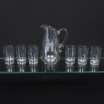 Conjunto em cristal diáfano e incolor, composto por uma(01) jarra e seis(06) copos. Jarra com corpo de perfil balaústre, bico vertedor pronunciado e alça lateral curvada aplicada sobre a superfície, copos de formato cilíndrico com base espessa, talhados com motivos geométricos estilizados em repetição vertical. Medidas: