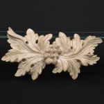 Talha de parede decorativa em resina moldada, representando composição vegetalista com folhas de acanto e cacho de frutos em relevo; modelagem em baixo e médio-relevo, acabamento em pátina clara simulando envelhecimento; verso plano com gancho para fixação. Medidas: