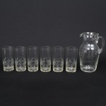 Seis(06) itens: a) Conjunto de copos em cristal, diáfano e incolor, bojo cilíndrico decorado por lapidação geométrica combinando motivos losangulares entrecruzados, com reservas em padrão quadriculado pontilhado e facetas angulares, dispostas em faixa contínua. Composto de seis(06) unidades. Medidas: ;b) Jarra em meio cristal incolor, corpo de perfil balaústre, talhado com sulcos que formam padrão de losangos e folhagens estilizadas, bico vertedor ondulado e revirado para fora, alça lateral curvada aplicada sobre a superfície. Medidas: